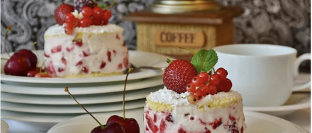 Zwei Dessertteller mit einem Kaffeemühle im Hintergrund. Ein Teller hat einen kleinen Kuchen mit Erdbeeren und roten Beeren oben, und der andere hat ein Dessert mit Kirschen und roten Beeren.