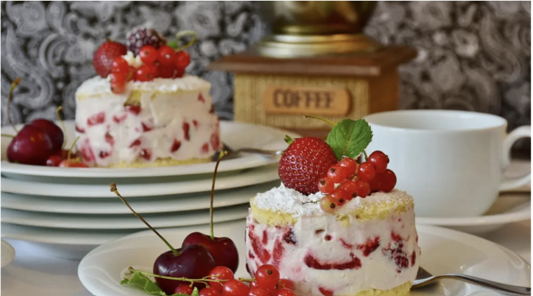 Zwei Dessertteller mit einem Kaffeemühle im Hintergrund. Ein Teller hat einen kleinen Kuchen mit Erdbeeren und roten Beeren oben, und der andere hat ein Dessert mit Kirschen und roten Beeren.