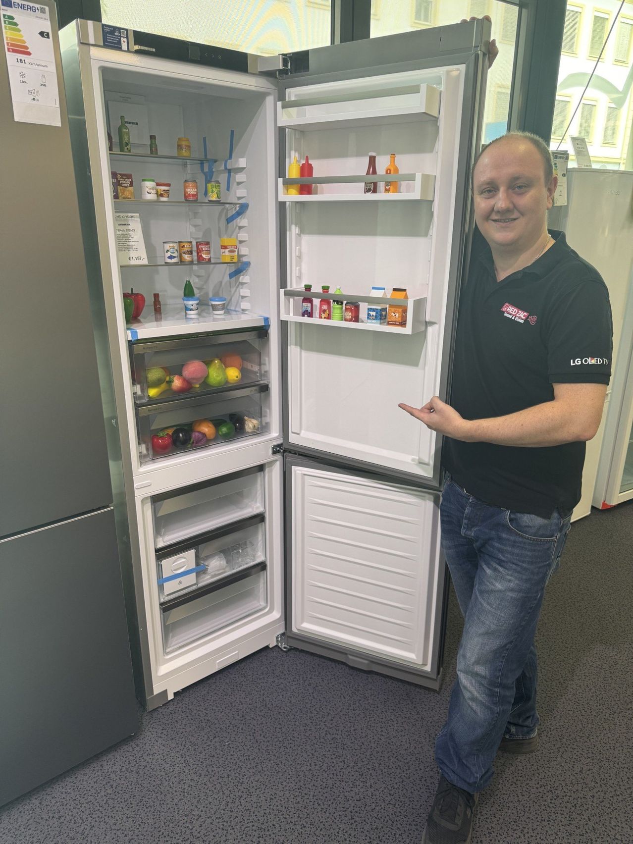 Ein Mann steht neben einem offenen Kühlschrank, der mit verschiedenen Lebensmitteln und Gewürzen gefüllt ist, er trägt ein schwarzes Poloshirt mit Logo.