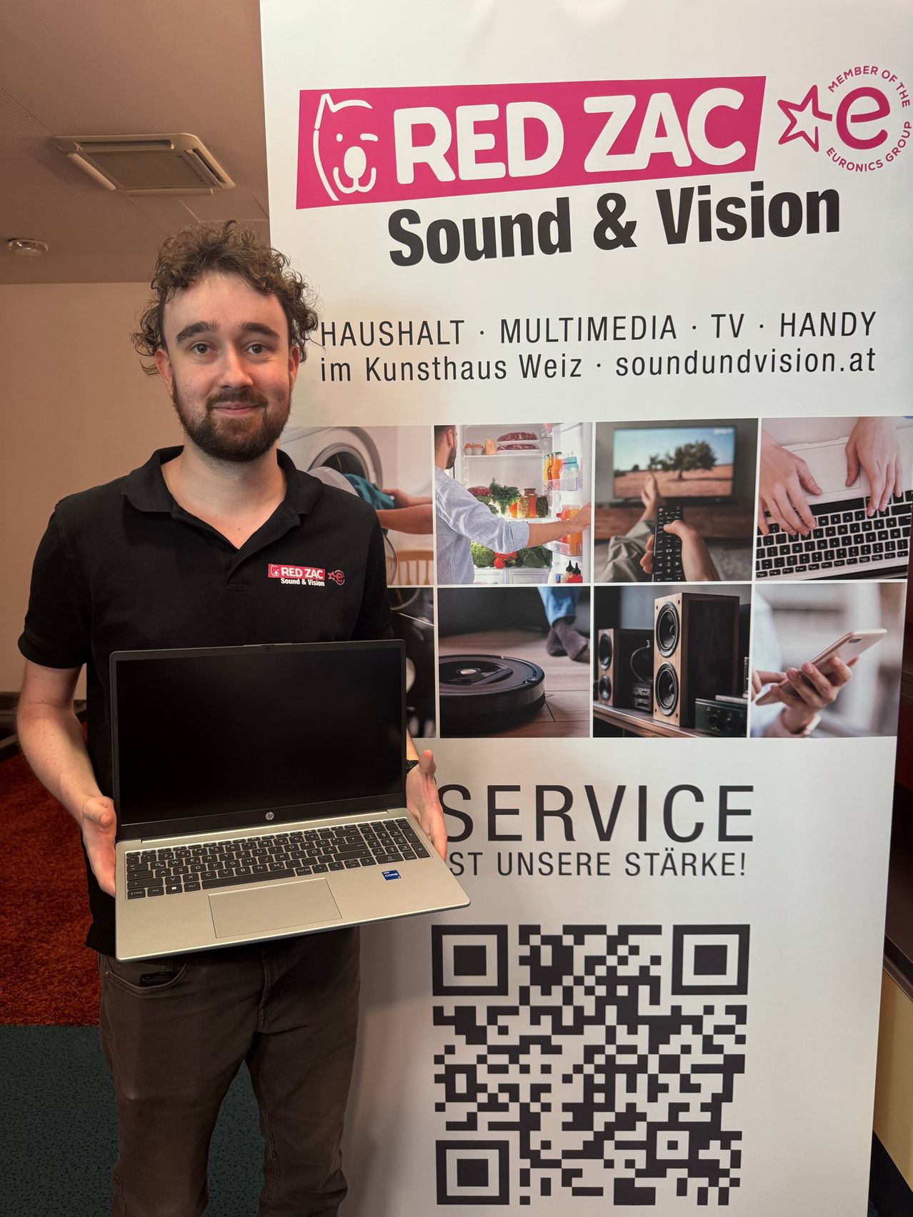 Ein Mann steht vor einem Plakat, das für Ton und Bild wirbt. Er hält einen Laptop in den Händen. Das Plakat zeigt einen QR-Code und den Slogan 'Service ist unsere Stärke!'.
