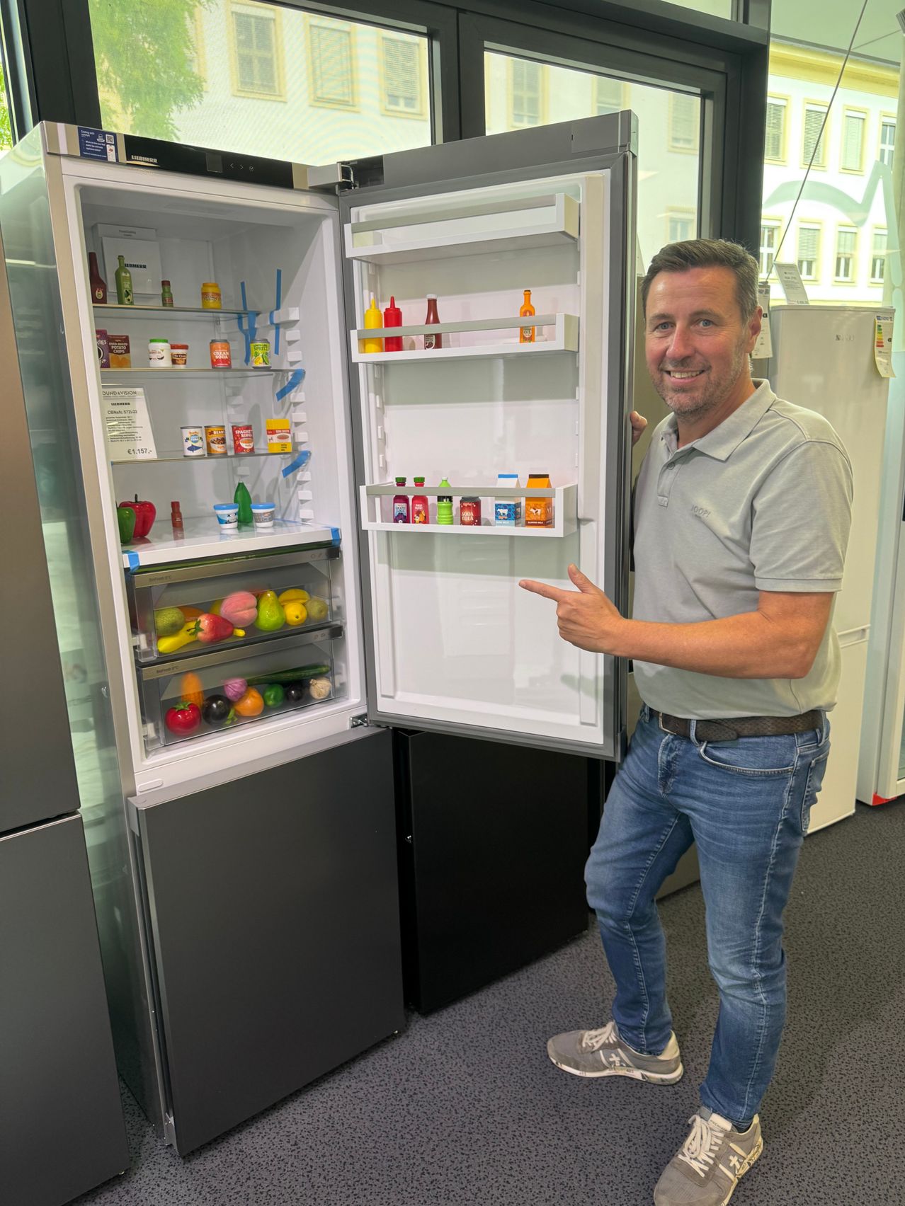Ein Mann steht neben einem offenen Kühlschrank, der mit Obst, Gemüse und Würzsaucen gefüllt ist. Er lächelt und zeigt auf den Kühlschrank.