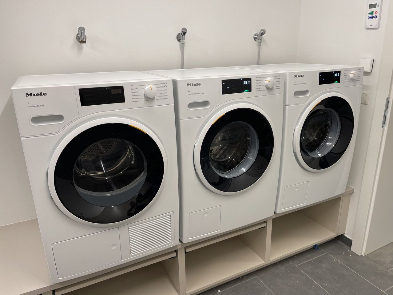 Drei Miele-Waschmaschinen sind auf einem weißen Regal gestapelt. Die Maschinen haben digitale Anzeigen, die die Zeit anzeigen. Die Wände und der Boden sind weiß und grau.