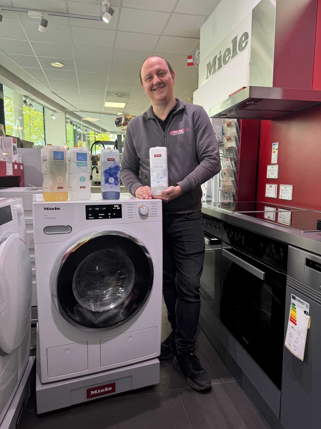 Ein Mann steht neben einer weißen Waschmaschine mit Waschmittelflaschen darauf und lächelt in einem Geschäft.