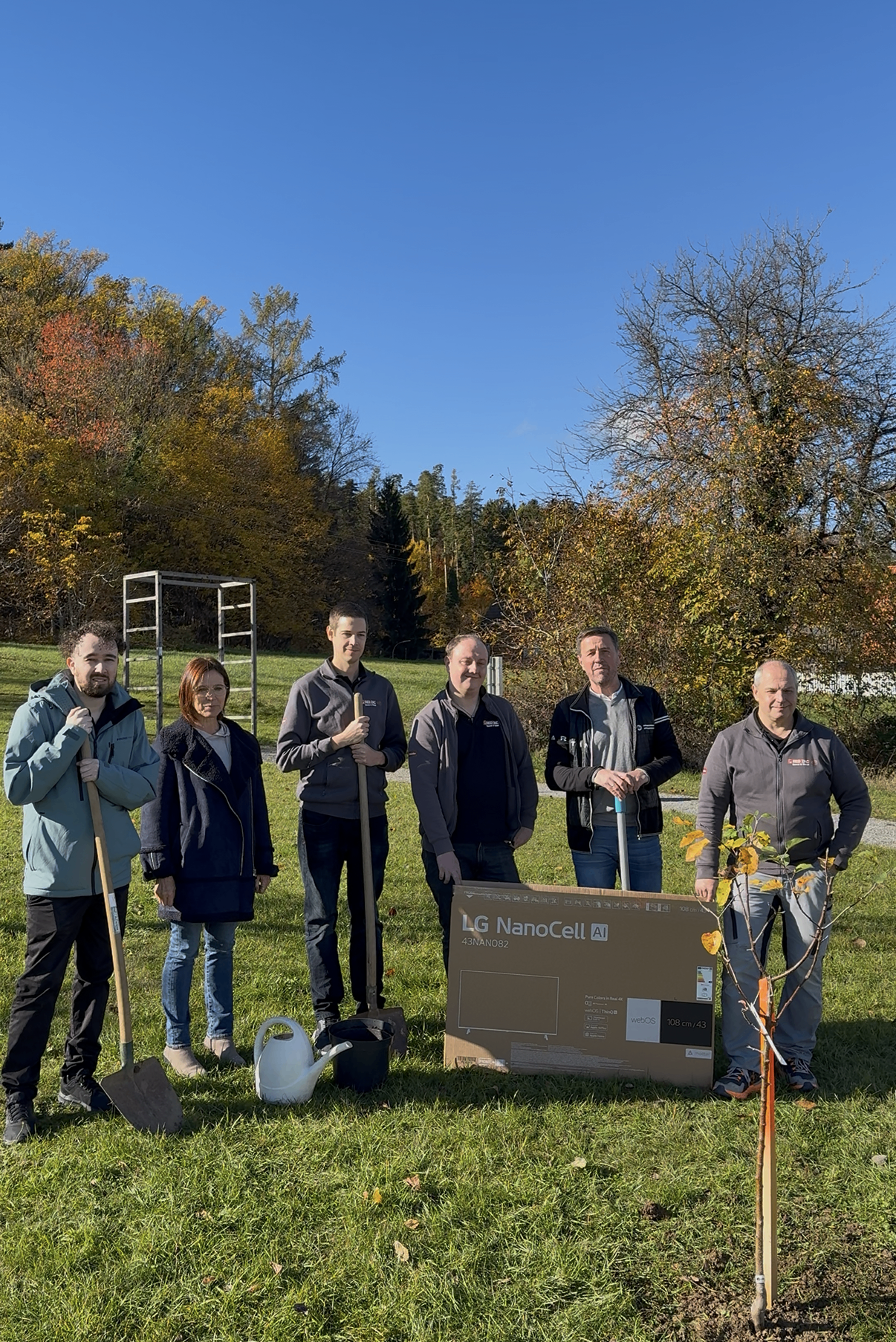 Sechs Personen stehen auf einem Rasen mit einer Schaufel und einem kleinen Baum. Ein Mann hält eine Gießkanne und eine Frau hält eine kleine Topfpflanze. Im Hintergrund sind Bäume mit bunten Herbstblättern zu sehen.