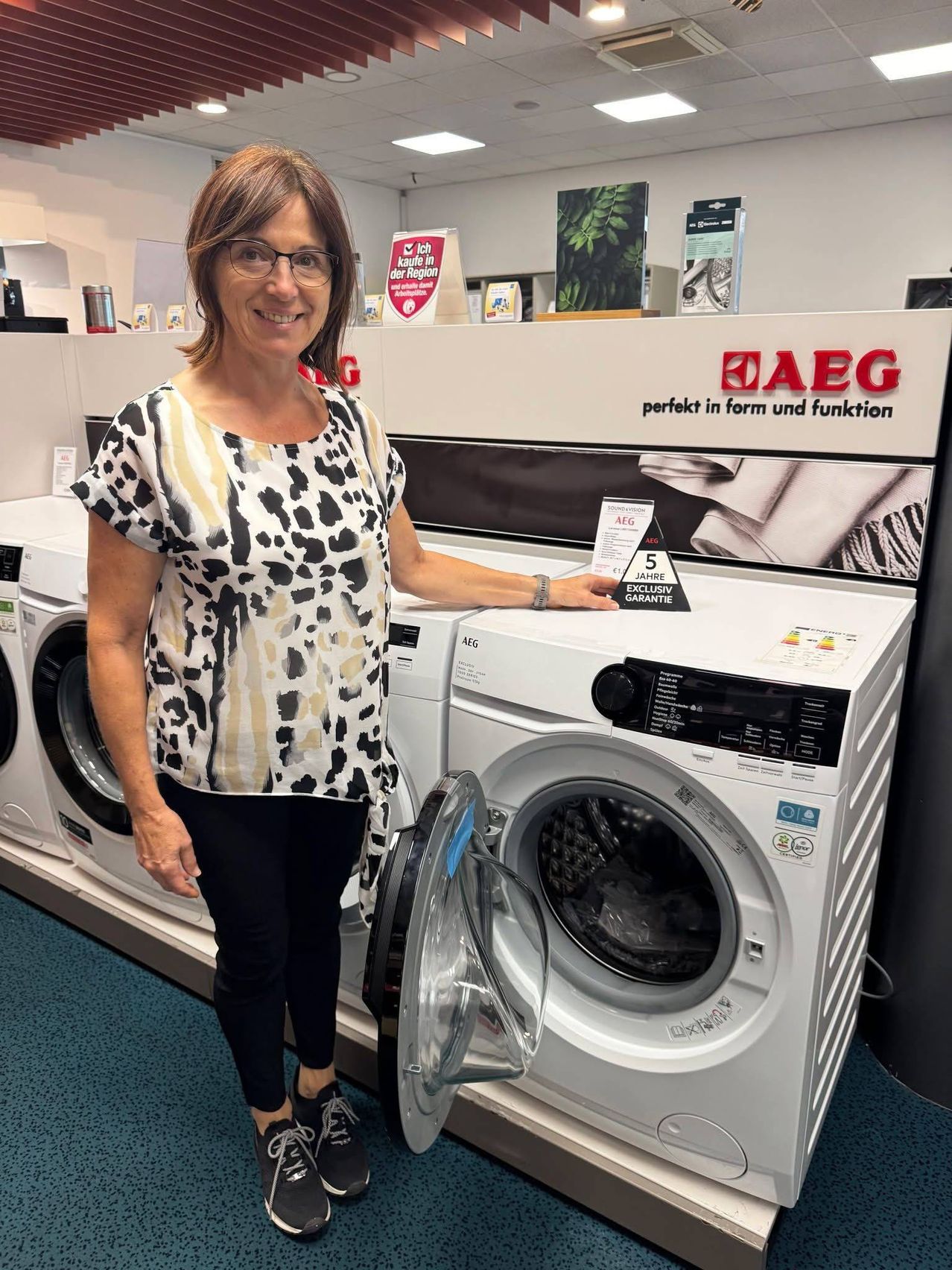 A woman stands in front of a white AEG washing machine with the door open, smiling for a photo. Behind her, there are more washing machines and a banner with the text 'perfect in form and function'.