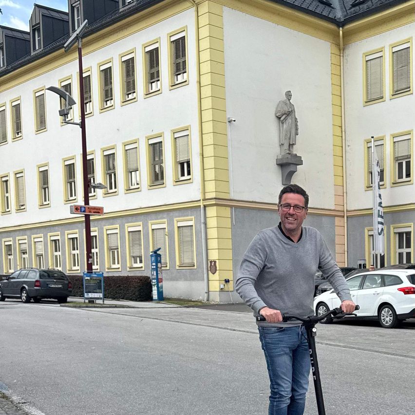 Ein Mann in einem grauen Pullover und Jeans fährt einen Roller auf einer Stadtstraße vor einem weißen Gebäude mit vielen Fenstern und einer Statue.