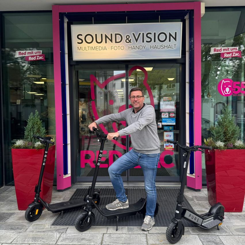 Ein Mann in einem grauen Pullover und blauen Jeans posiert auf zwei schwarzen Elektrorollern vor einem Laden namens Sound & Vision.