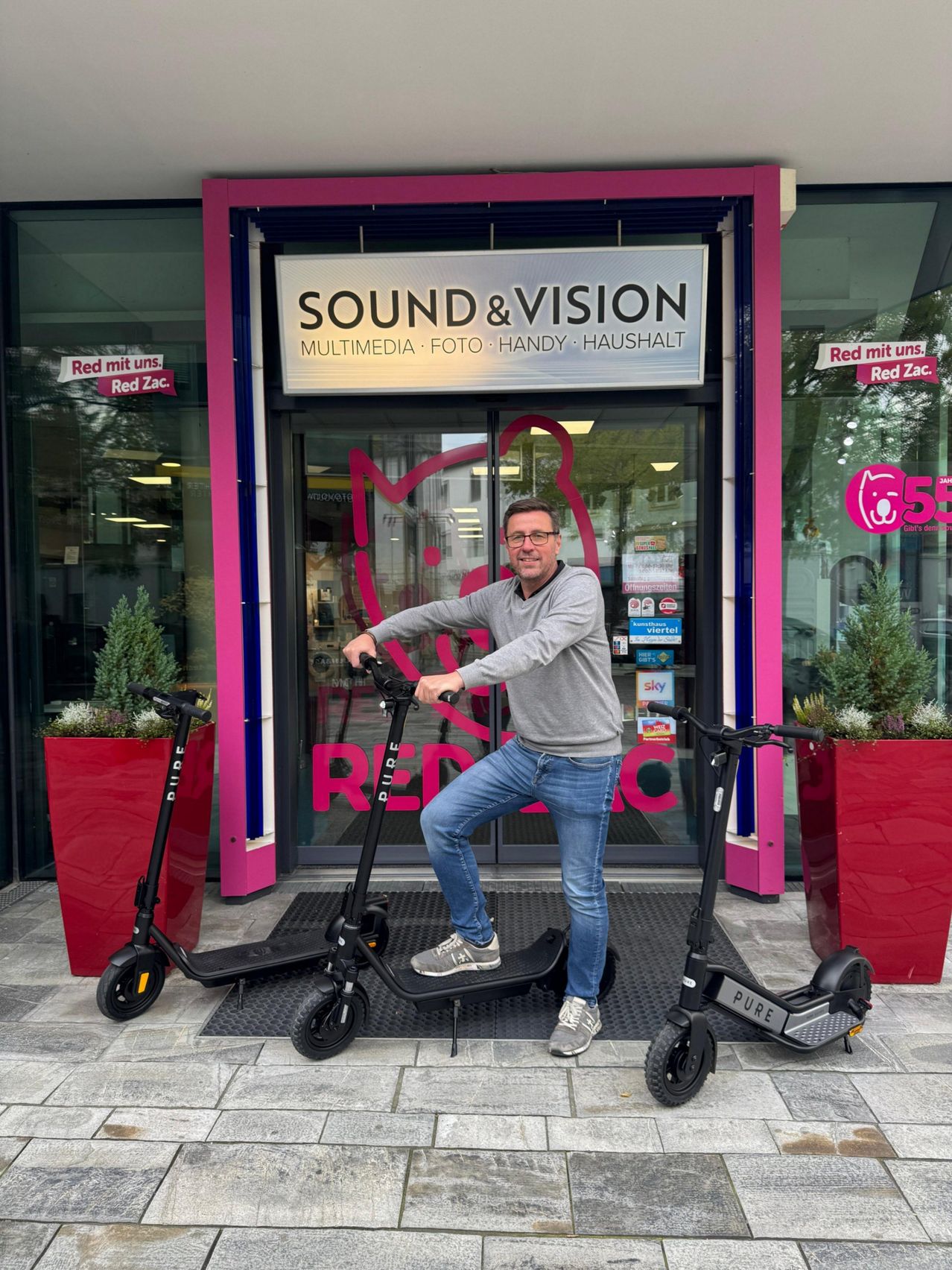 Ein Mann mit Brille und grauem Pullover steht auf einem schwarzen Teppich vor einem Geschäft mit dem Schild 'Sound & Vision'. Er hält einen schwarzen Roller mit dem Wort 'Pure' darauf. Vor ihm stehen zwei Roller und zwei Topfpflanzen auf beiden Seiten des Eingangs.