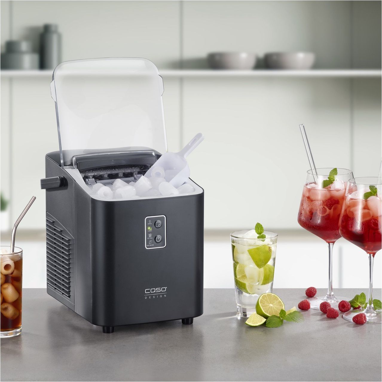 Eine schwarze Eismaschine mit durchsichtigem Deckel steht auf einer Arbeitsplatte. Sie hat einen Eislöffel und Eiswürfel im Inneren. Auf der Arbeitsplatte befinden sich drei Gläser mit Getränken und Garnierungen. Ein Glas hat eine Limettenscheibe und Minzblätter. Ein weiteres Glas hat eine Himbeere und ein Minzblatt. Das dritte Glas hat eine Erdbeere und ein Minzblatt.