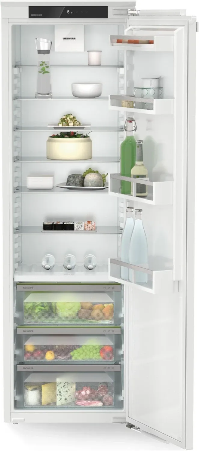 An open refrigerator with shelves holding a variety of food items, including a cake, cheese, and vegetables. The bottom drawer contains more vegetables.