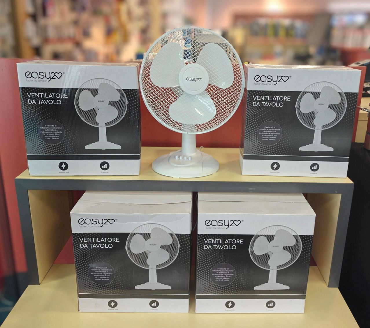 Drei weiße und schwarze Kartons eines Tischventilators sind auf einem Holzregal gestapelt. Die mittlere Schachtel enthält einen echten Tischventilator.