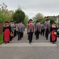 Eine Blaskapelle in traditioneller Kleidung geht einen Bürgersteig entlang. Sie tragen Instrumente wie Trompeten und Posaunen.