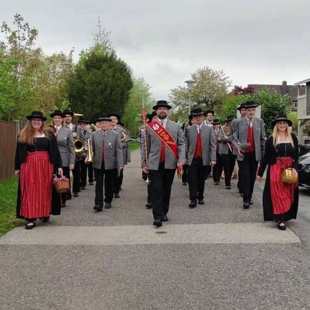Eine Blaskapelle in traditioneller Kleidung geht einen Bürgersteig entlang. Sie tragen Instrumente wie Trompeten und Posaunen.