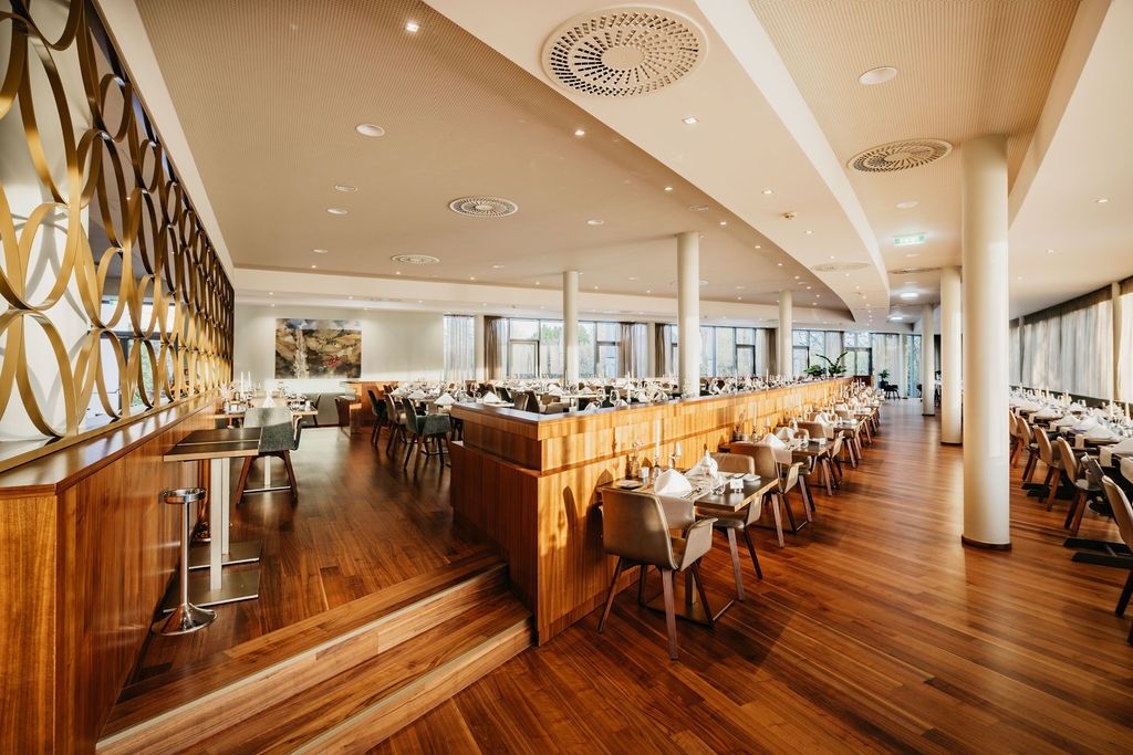 Ein elegantes Restaurant mit Holzböden und hohen Fenstern. Die Tische sind mit weißen Tüchern und Kerzen gedeckt. Eine lange Holztheke mit Stühlen verläuft entlang der Wand.