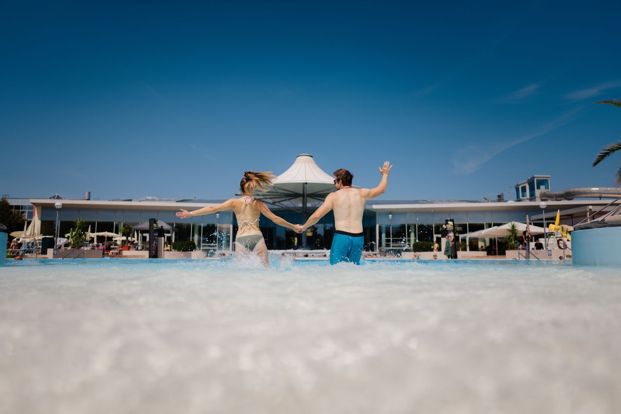 Ein Paar in Badekleidung, das in einem Pool tanzt und spritzt, mit einem modernen Gebäude im Hintergrund.