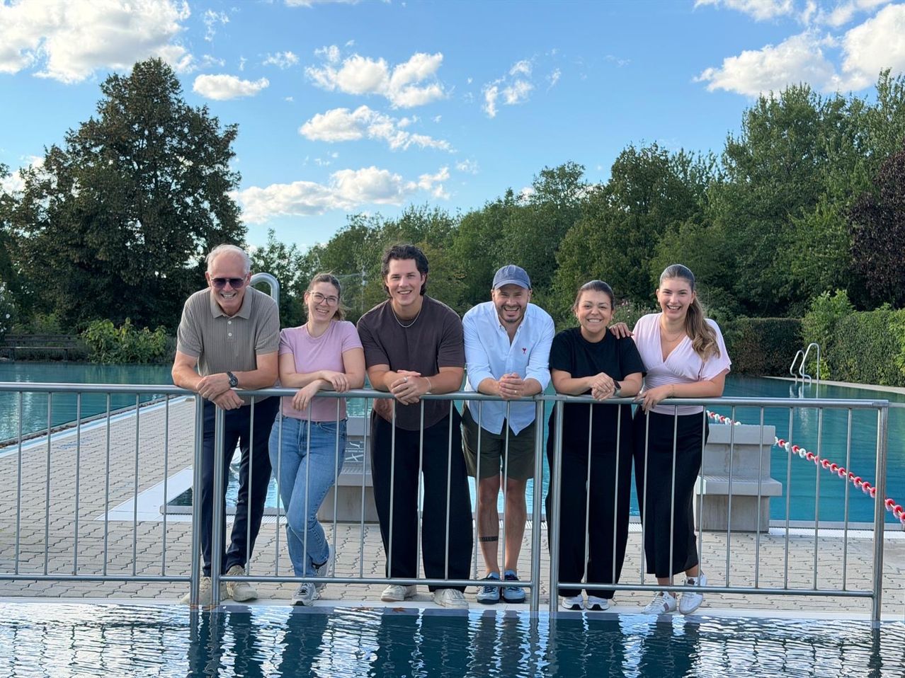 A group of six people standing by a pool, smiling and posing for a photo. They are leaning on a metal fence. Behind them, there is a pool with a concrete deck. Trees and a blue sky with clouds are in the background.