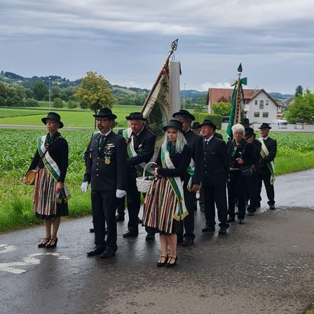 Eine Gruppe von Menschen in traditioneller Kleidung, darunter zwei Frauen, marschiert in einer Reihe auf einem Feld. Sie tragen Fahnen und Körbe. Der Himmel ist bewölkt.