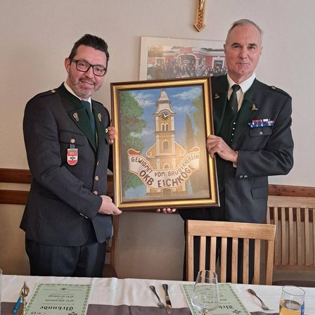 Zwei Männer in Uniformen halten ein gerahmtes Gemälde einer Kirche mit einer deutschen Inschrift. Sie stehen in einem Esszimmer mit Stühlen und einem Tisch, mit einem Kreuz an der Wand hinter ihnen.