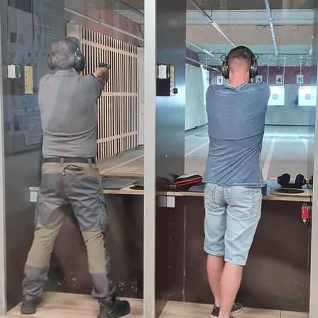 Zwei Männer in einem Schießstand. Einer trägt ein graues Shirt, Hose und Kopfhörer. Der andere trägt ein blaues Shirt, Jeansshorts und Kopfhörer. Beide zielen mit ihren Waffen auf Ziele.