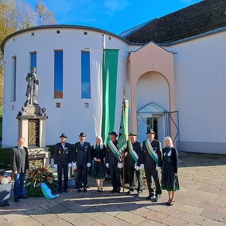 Eine Gruppe von Personen steht vor einem Denkmal und hält Fahnen. Dahinter befindet sich eine Statue und ein Gebäude mit Glasfenstern.
