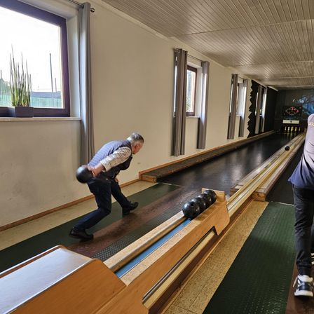 Ein älterer Mann in einer Bowlingbahn wirft einen Bowlingball, während eine weitere Person hinter der Bahn steht.