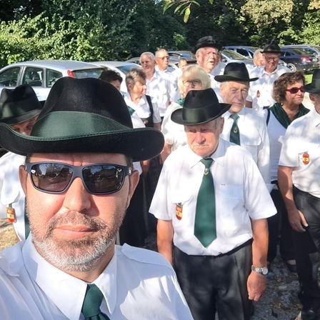 Eine Gruppe von Menschen in weißen Uniformen und grünen Krawatten, mit Hüten, steht in einem Park, mit geparkten Autos dahinter.