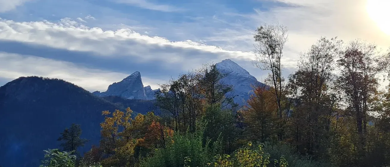 Ein Blick auf eine Berglandschaft mit schneebedeckten Gipfeln und herbstlichen Bäumen unter einem blauen Himmel mit Wolken.