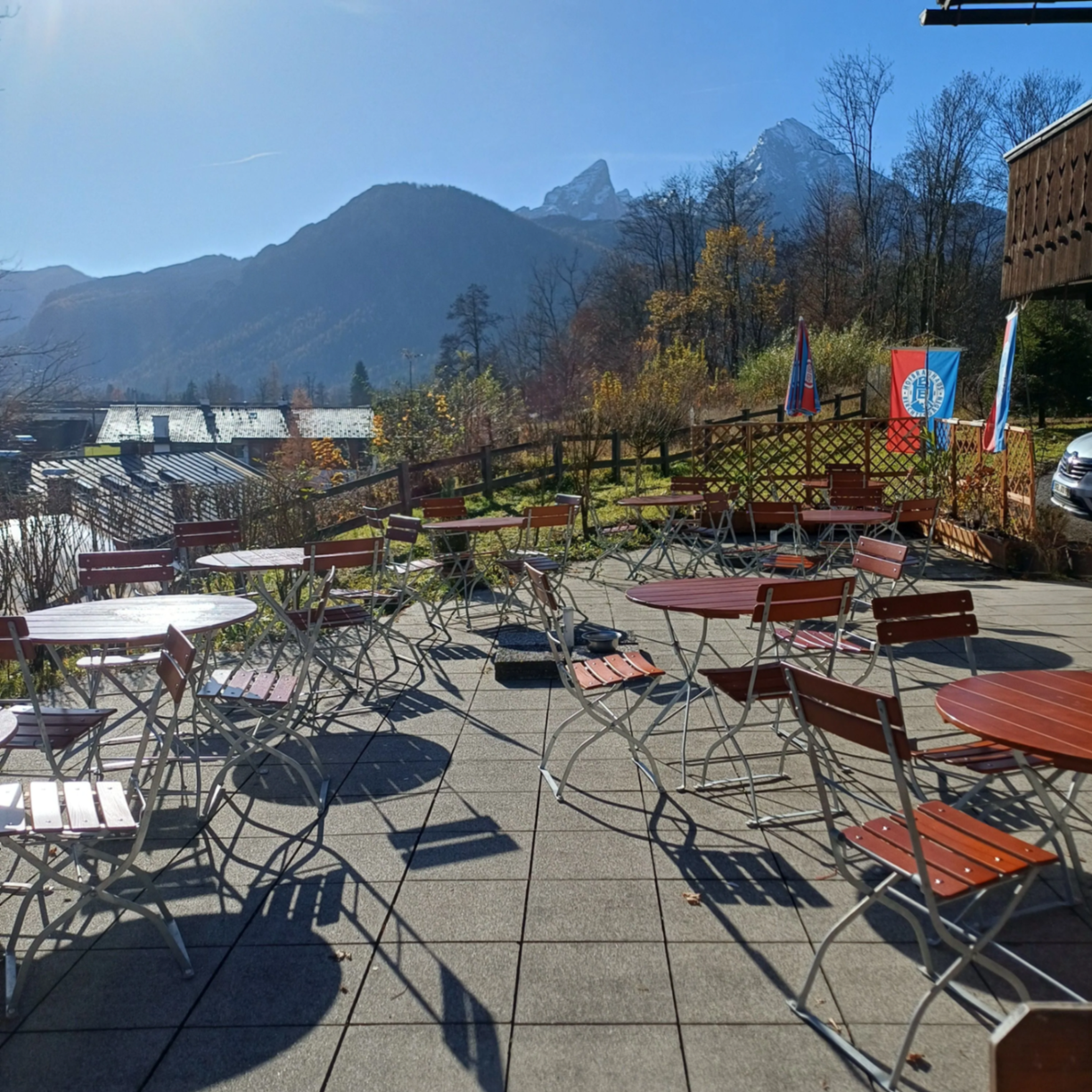 Terrasse eines Restaurants mit Tischen und Stühlen, Blick auf die Berge. Ein klarer Himmel und mehrere Fahnen sind sichtbar.
