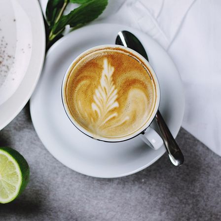 Eine Tasse Kaffee mit Blattdesign steht auf einer Untertasse, neben einem Teller mit Kuchen und Limetten.