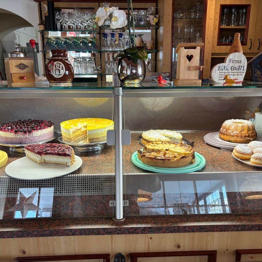 Eine Bäckerei-Vitrine mit verschiedenen Kuchen und Gebäck, darunter Käsekuchen und ein Gugelhupf, vor dem Hintergrund von Regalen mit Glaswaren und Dekorationsartikeln.