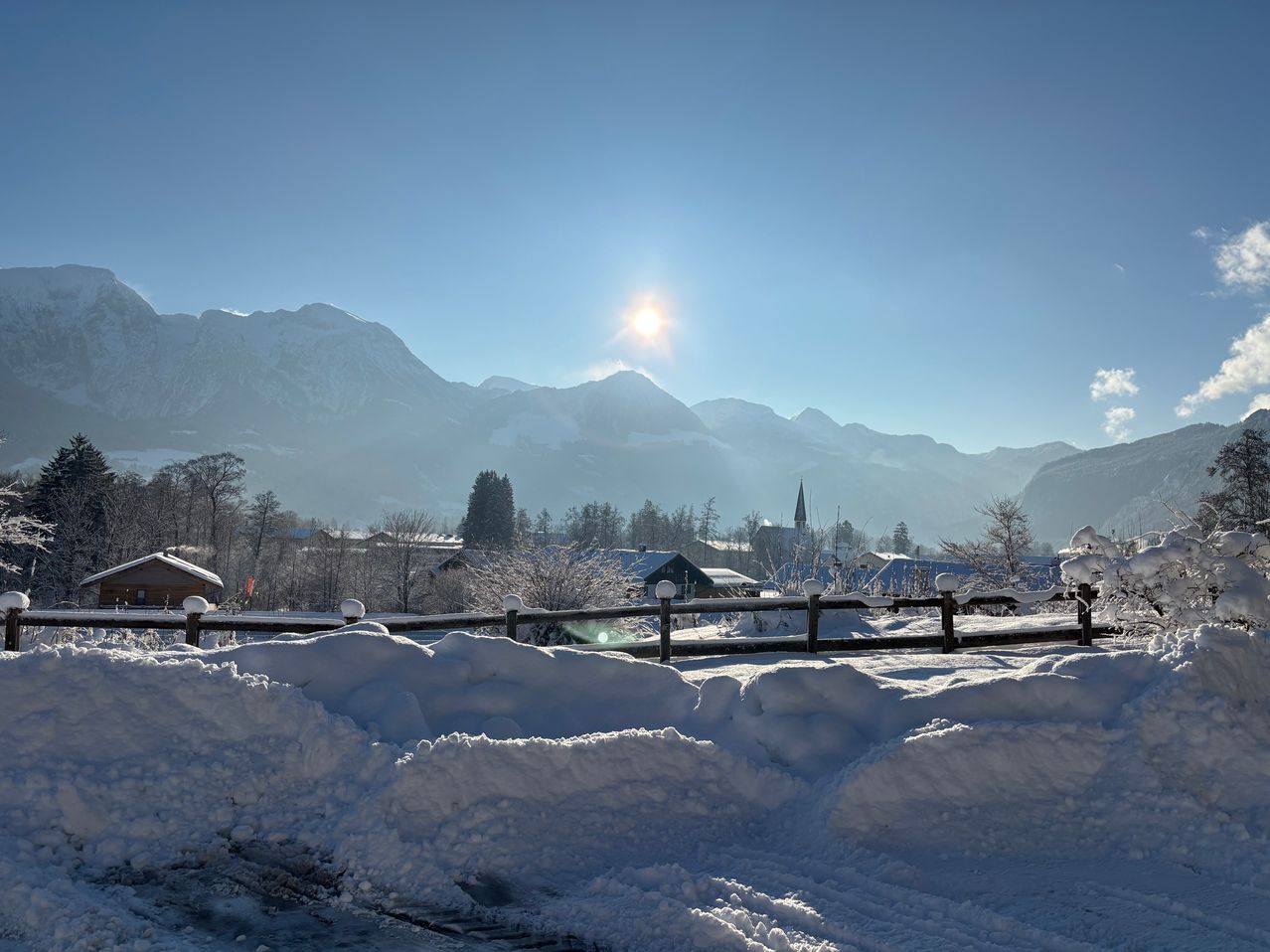 Eine verschneite Landschaft mit Bergen, Bäumen und Häusern, die mit Schnee bedeckt sind, und einer hellen Sonne, die durch die Wolken scheint.