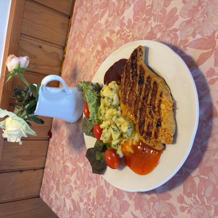Ein weißer Teller mit gegrilltem Hähnchen, Kartoffelpüree und Salat mit roten Tomaten und weißen Rosen auf einer Blumentischdecke.