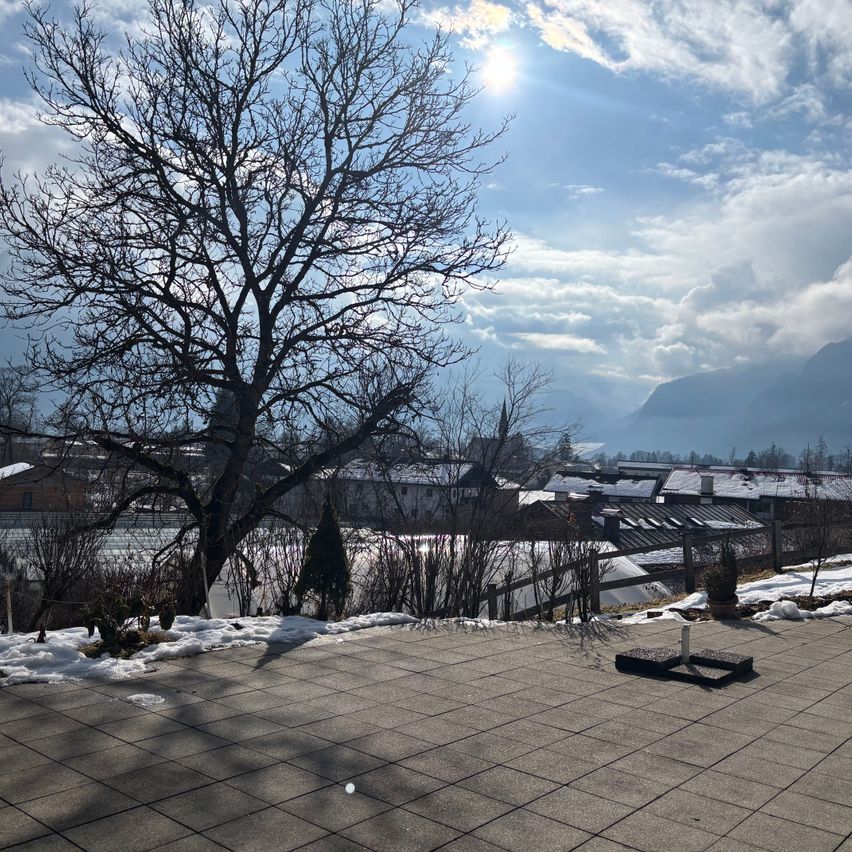 Ein Blick auf die Stadtlandschaft von einer Dachterrasse, mit einem laublosen Baum, Schnee auf dem Boden und einer strahlenden Sonne am teilweise bewölkten Himmel mit Bergen in der Ferne.