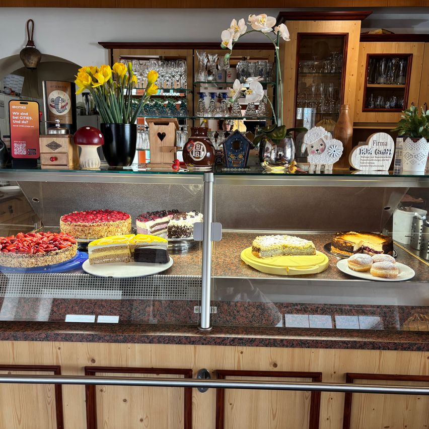 Ein Tresen voller verschiedener Kuchen und Gebäck, einschließlich mehrschichtiger Kuchen mit Früchten und Schokolade sowie Donuts, ausgestellt unter einer Glasabdeckung. Der Tresen hat Holzschränke und Regale im Hintergrund.