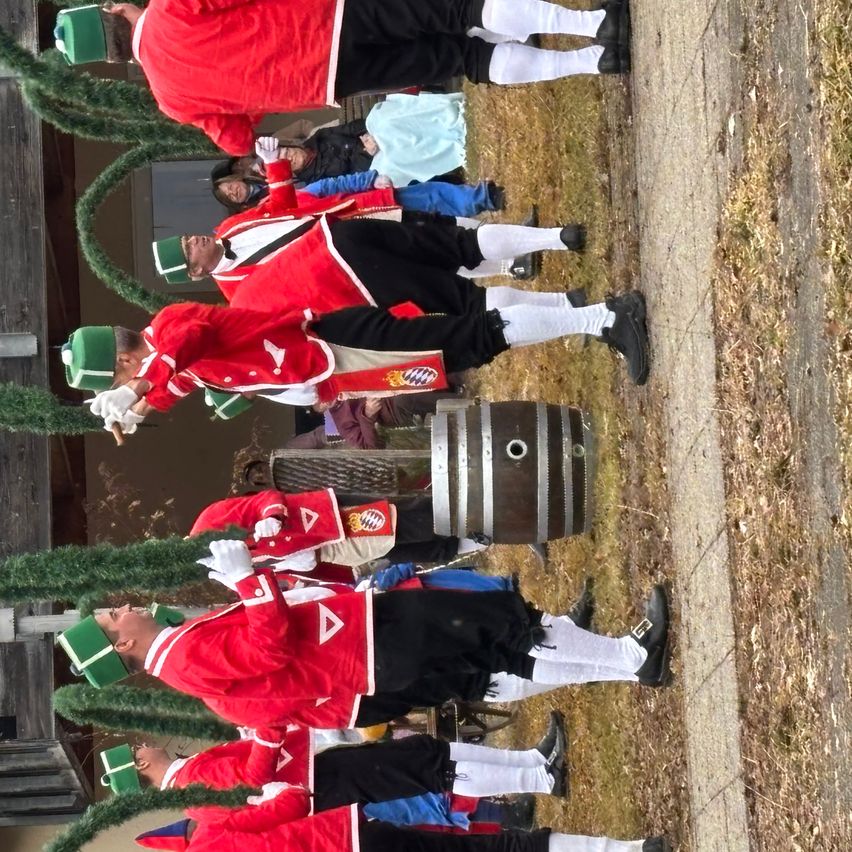 Eine Gruppe von Personen, die rote und schwarze Uniformen mit grünen Mützen tragen, steht auf Gras, möglicherweise bei einer Aufführung. Eine Person hält einen Fass. Im Hintergrund sind weitere Personen in unterschiedlicher Kleidung zu sehen.