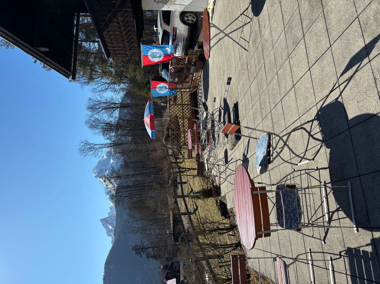 Eine Ansicht eines Außencafés mit Klapptischen und Stühlen. Im Vordergrund sind Fahnen zu sehen, und im Hintergrund befinden sich schneebedeckte Berge.