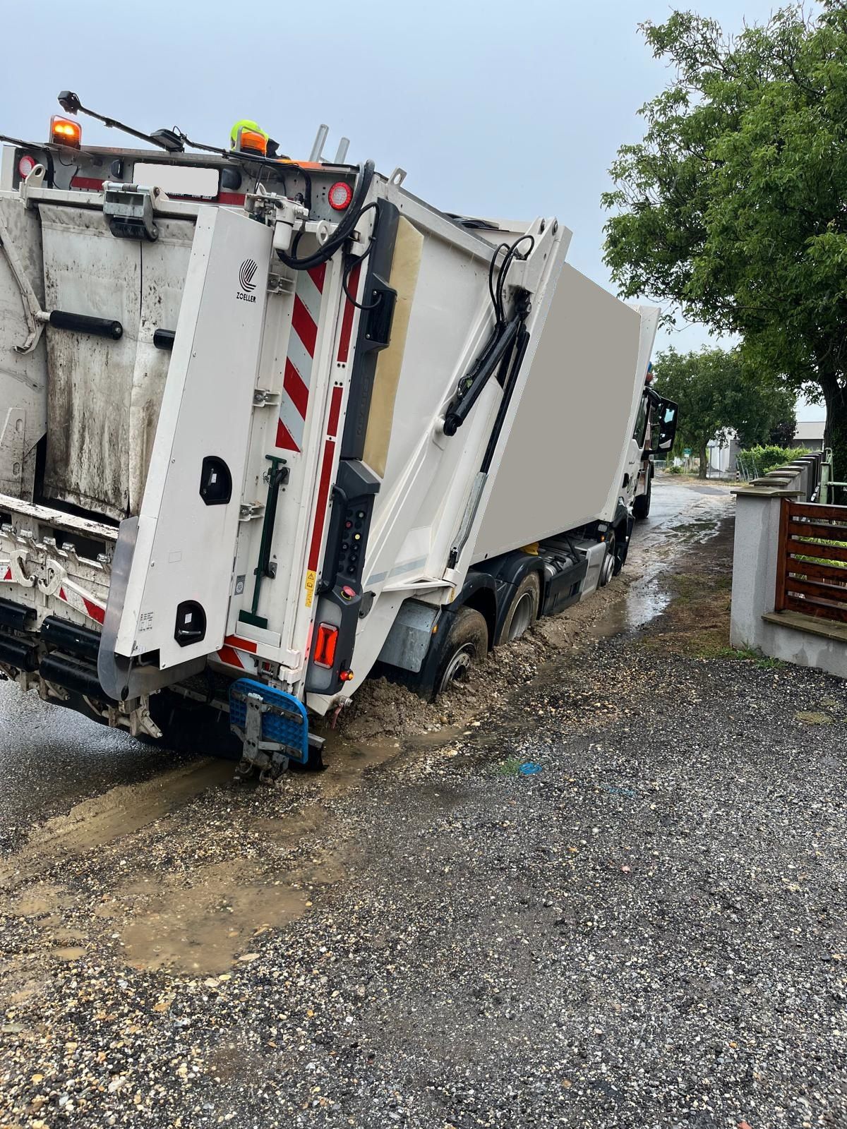 Ein Müllwagen steckt im Schlamm fest, wobei die hintere Tür offen ist und der Container zur Seite geneigt ist. Bäume und ein Zaun sind im Hintergrund sichtbar.