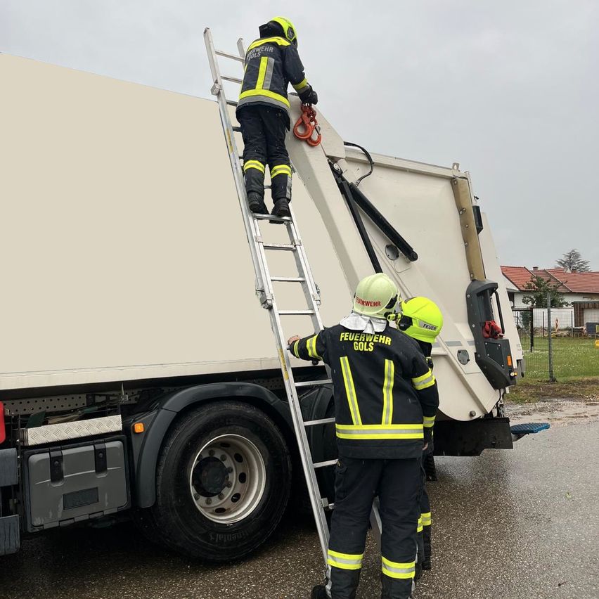 Drei Feuerwehrleute in Uniform klettern auf eine Leiter an der Seite eines Lastwagens. Der Lastwagen ist weiß mit schwarzen Reifen. Einer der Feuerwehrleute hält sich an einem Haken fest.