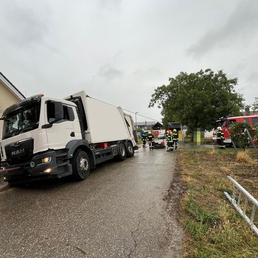 Ein weißer MAN-Lkw ist auf einer Straße geparkt, mit Feuerwehrleuten in gelben Helmen in der Nähe. Ein roter Feuerwehrwagen ist im Hintergrund geparkt.