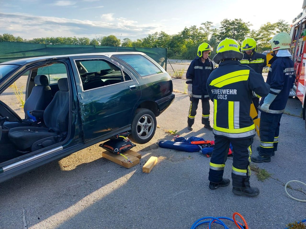 Drei Feuerwehrleute stehen bei einem beschädigten Auto, eines davon stützt es mit einem Wagenheber. Die Autotür ist offen, und sie haben Sicherheitsausrüstung in der Nähe.