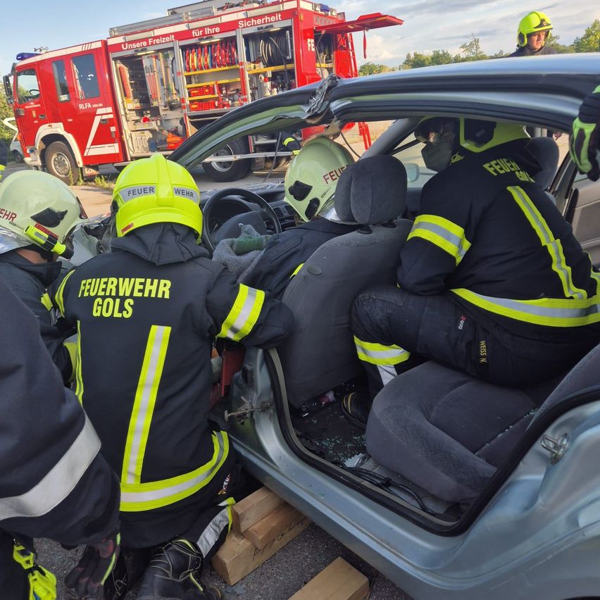 Feuerwehrleute in Schutzausrüstung helfen Personen aus einem beschädigten Auto. Die Tür des Fahrzeugs ist geöffnet, und ein Feuerwehrauto ist in der Nähe geparkt.