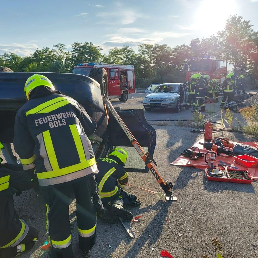 Rettungskräfte in gelben Uniformen arbeiten am Unfallort eines Fahrzeugs, mit Werkzeugen und Ausrüstung auf dem Boden verteilt. Ein Auto ist umgekippt, und Feuerwehrwagen sind in der Nähe geparkt.