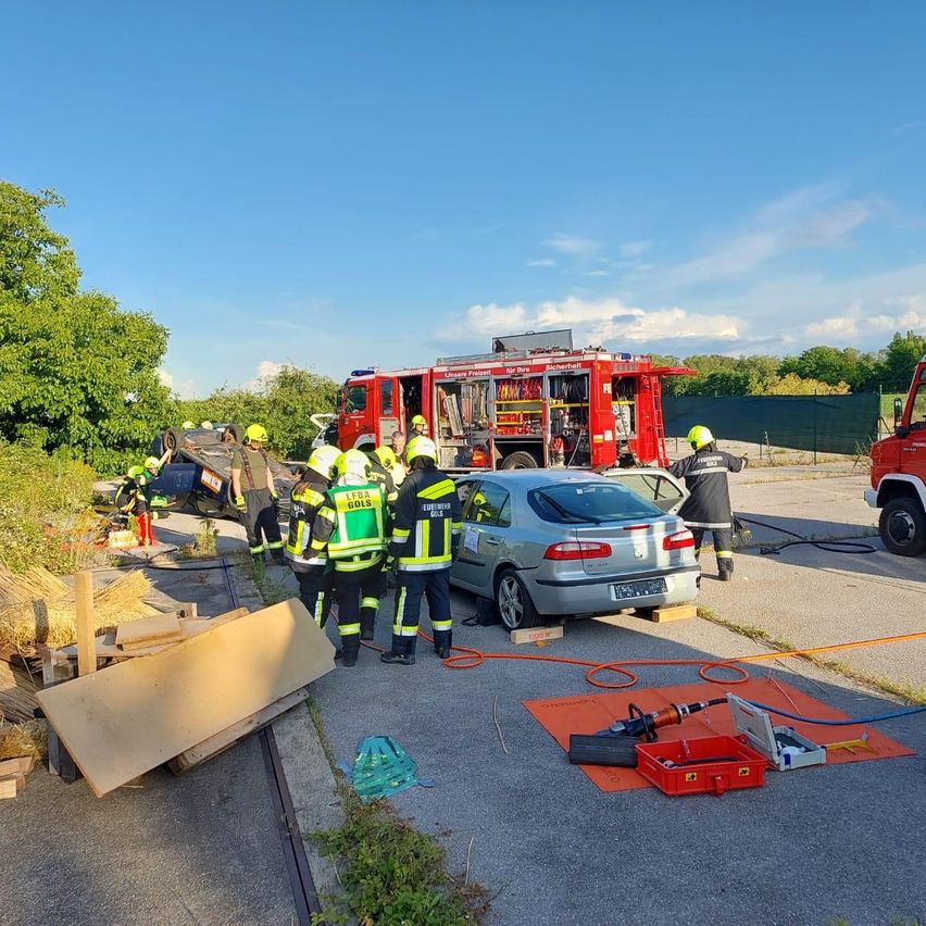 Eine Gruppe Feuerwehrleute arbeitet an einem Unfallort, einer von ihnen in der Nähe eines Autos und andere in der Nähe eines roten Feuerwehrwagens.