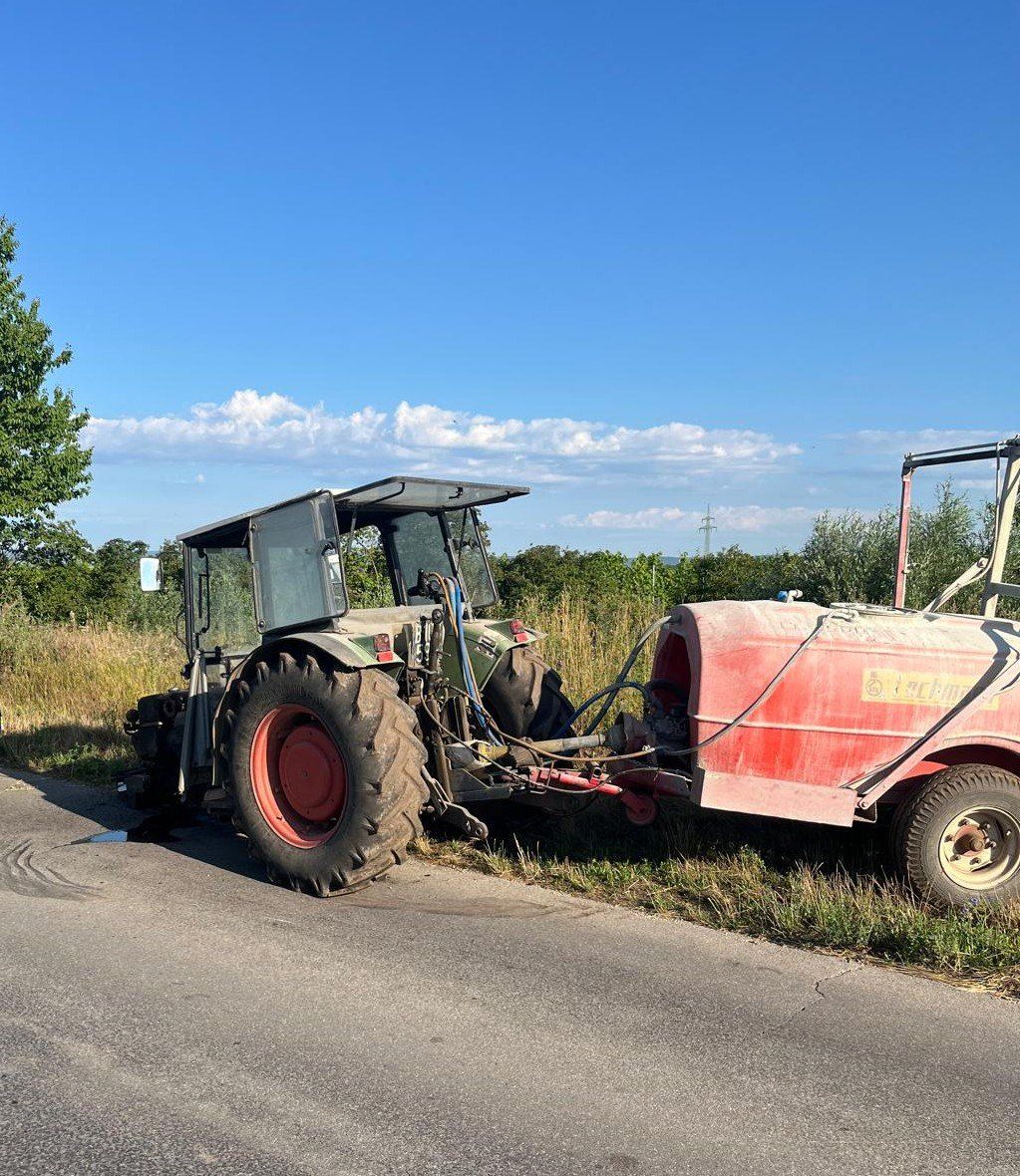 Ein roter Traktor mit Anhänger steht am Straßenrand. Der Traktor hat ein großes rotes Rad und sprüht Wasser aus dem Anhänger.