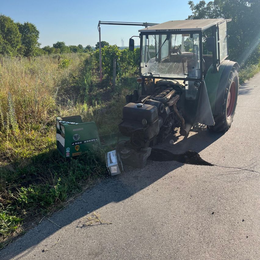 Ein alter Traktor ist am Straßenrand geparkt. Er hat eine zerbrochene Windschutzscheibe und einen beschädigten vorderen Stoßfänger. In der Nähe befindet sich ein grüner Kasten mit Text und einem Aufkleber.
