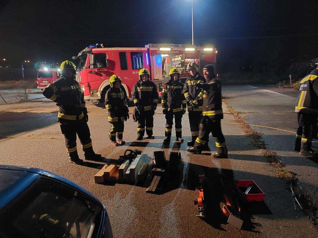 A group of firefighters is gathered at night near a fire truck, with tools and equipment scattered on the ground.