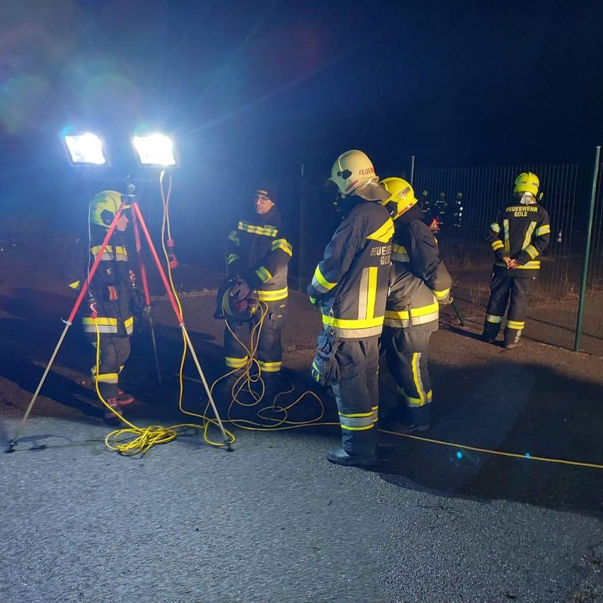 A group of firefighters in uniform stand outdoors at night. One holds a tool, another is adjusting a tripod with lights. They are illuminated by a bright light source.