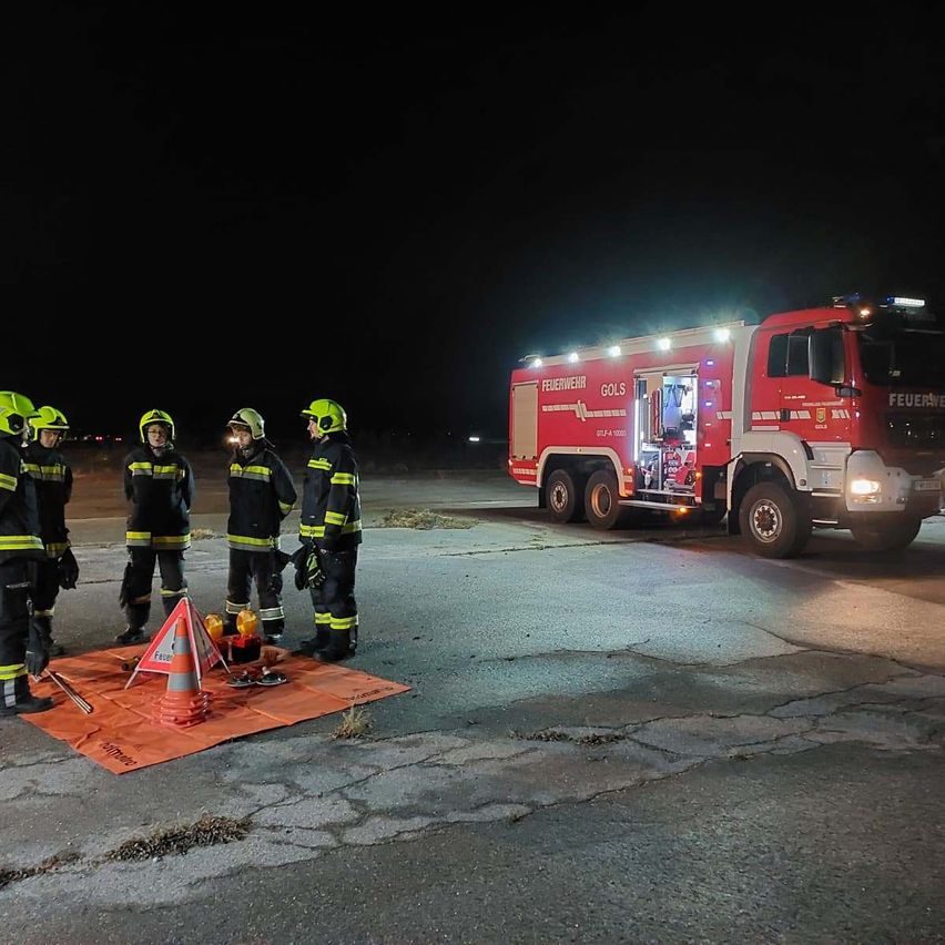 Five firefighters are gathered in a nighttime setting, with a fire truck parked behind them. They are equipped with yellow helmets and protective gear.