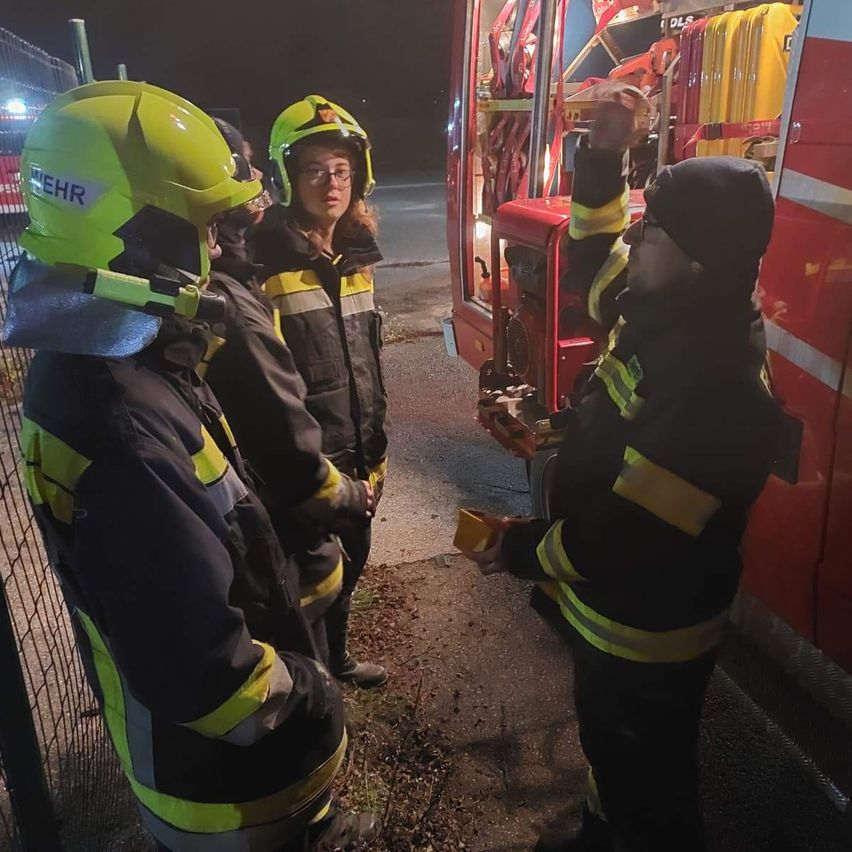 Firefighters in yellow helmets and jackets are gathered around a fire truck. One person is pointing at the truck while others watch.