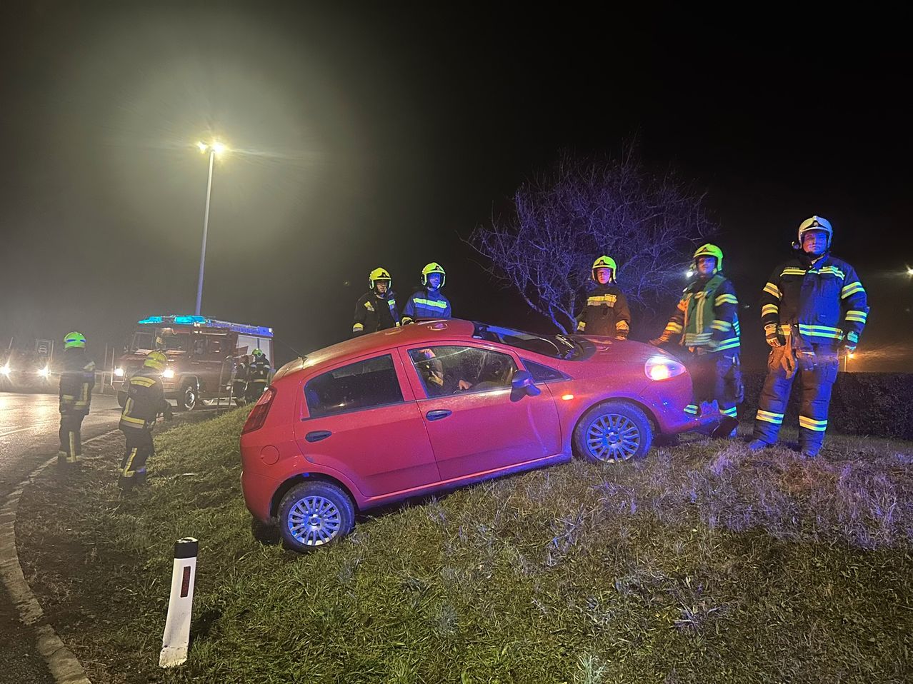 Ein rotes Auto ist nachts am Straßenrand kaputt. Notfallhelfer in gelber und grüner Kleidung beurteilen die Situation.